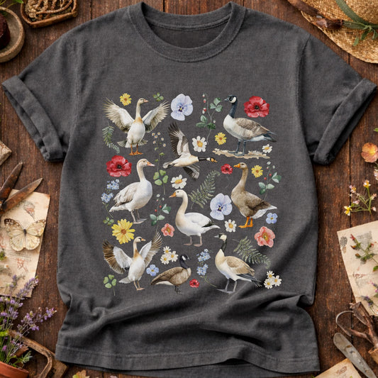 A dark gray crewneck t-shirt featuring a detailed graphic print of various waterfowl birds and colorful wildflowers, laid flat on a rustic wooden surface surrounded by gardening and vintage-style props.